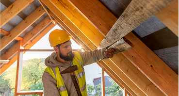 Construction worker in hard hat and safety vest installing insulation in wooden roof rafters