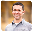 Professional headshot of smiling man in gray collared shirt against blurred background