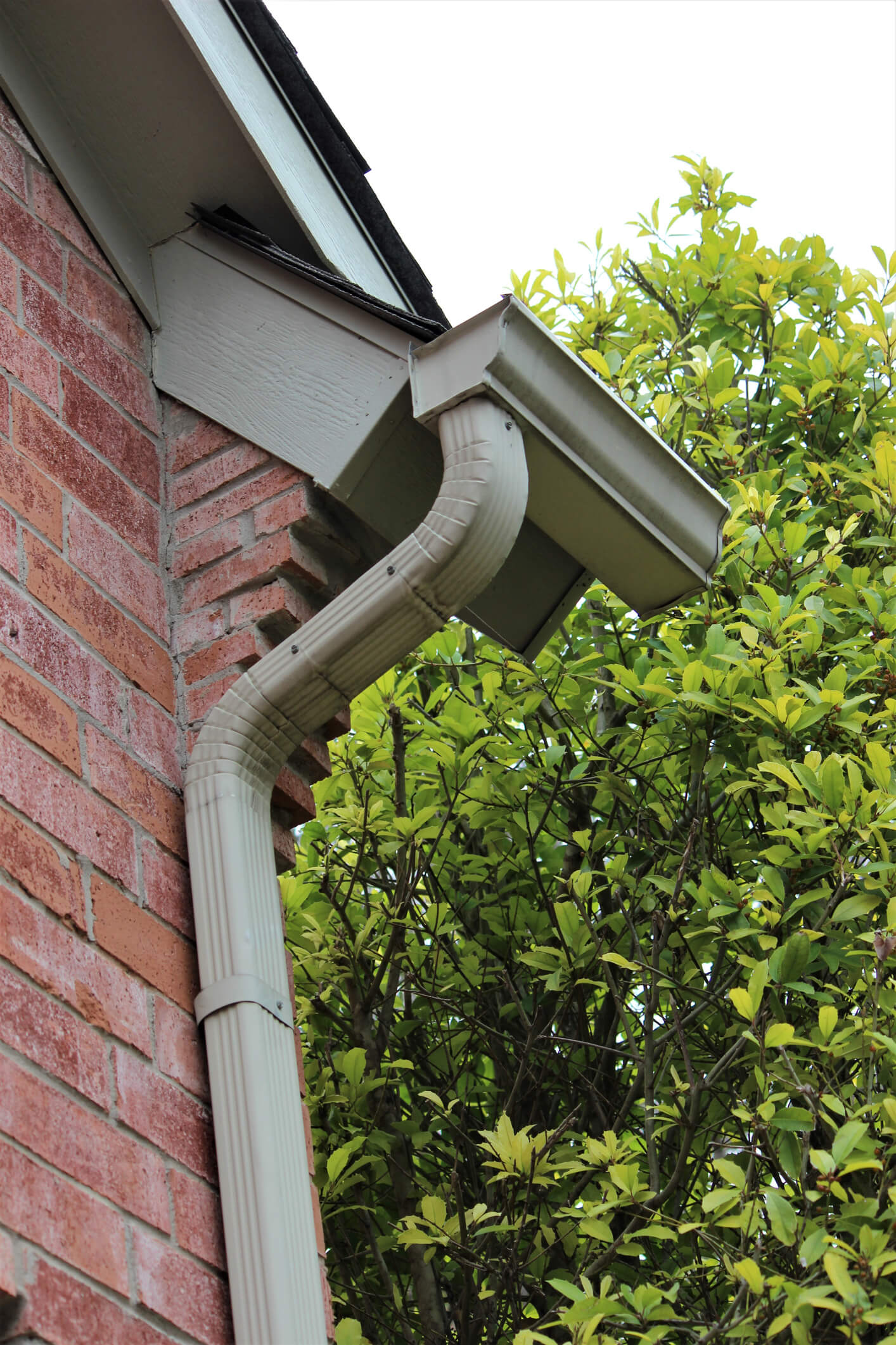 White rain gutter and downspout system on brick house exterior with green foliage