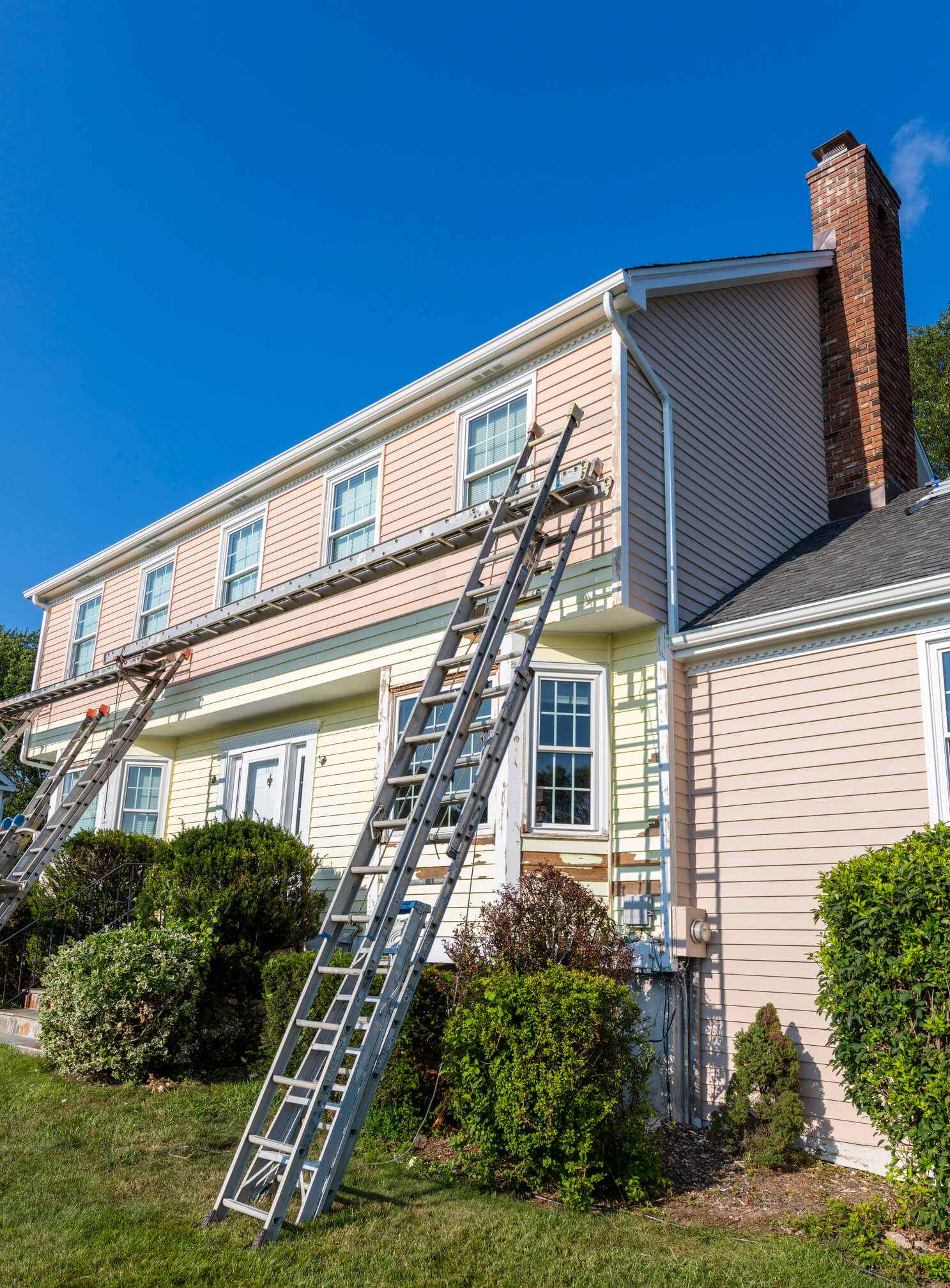 Two-story colonial house with siding being painted, extension ladders against exterior wall