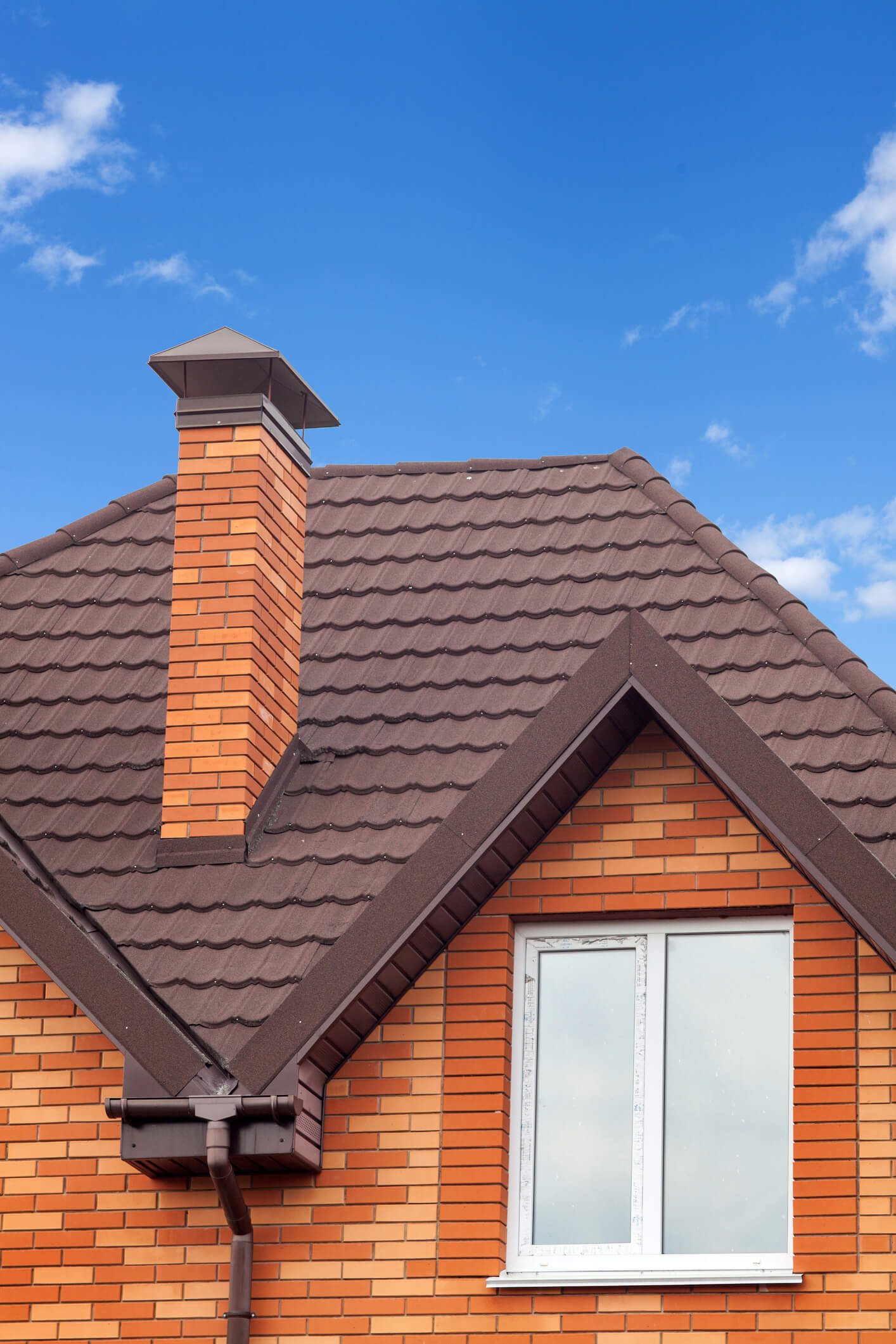 Orange brick house with brown tile roof and chimney against blue sky with clouds