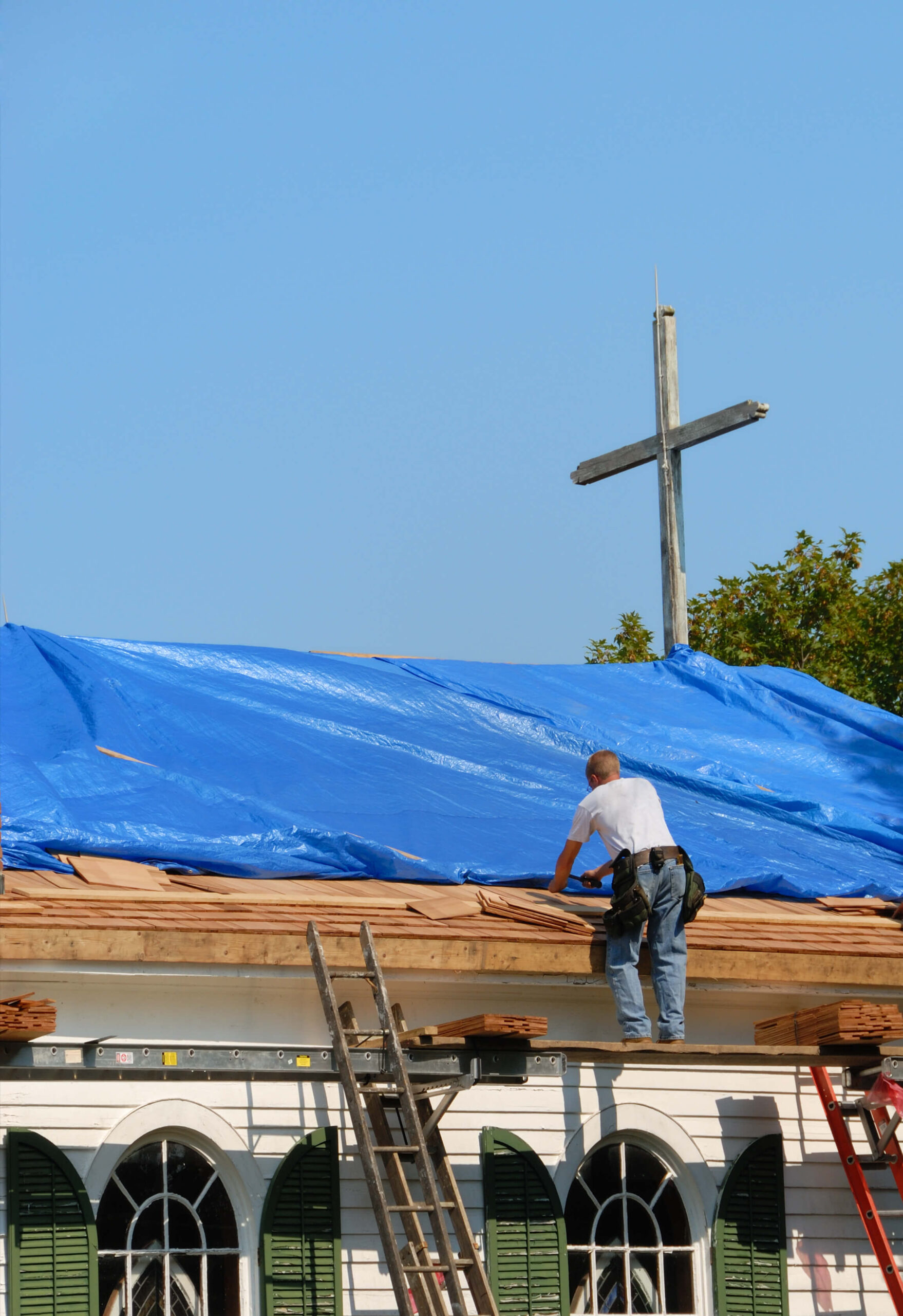 Construction worker installing blue tarp on church roof with wooden cross visible
