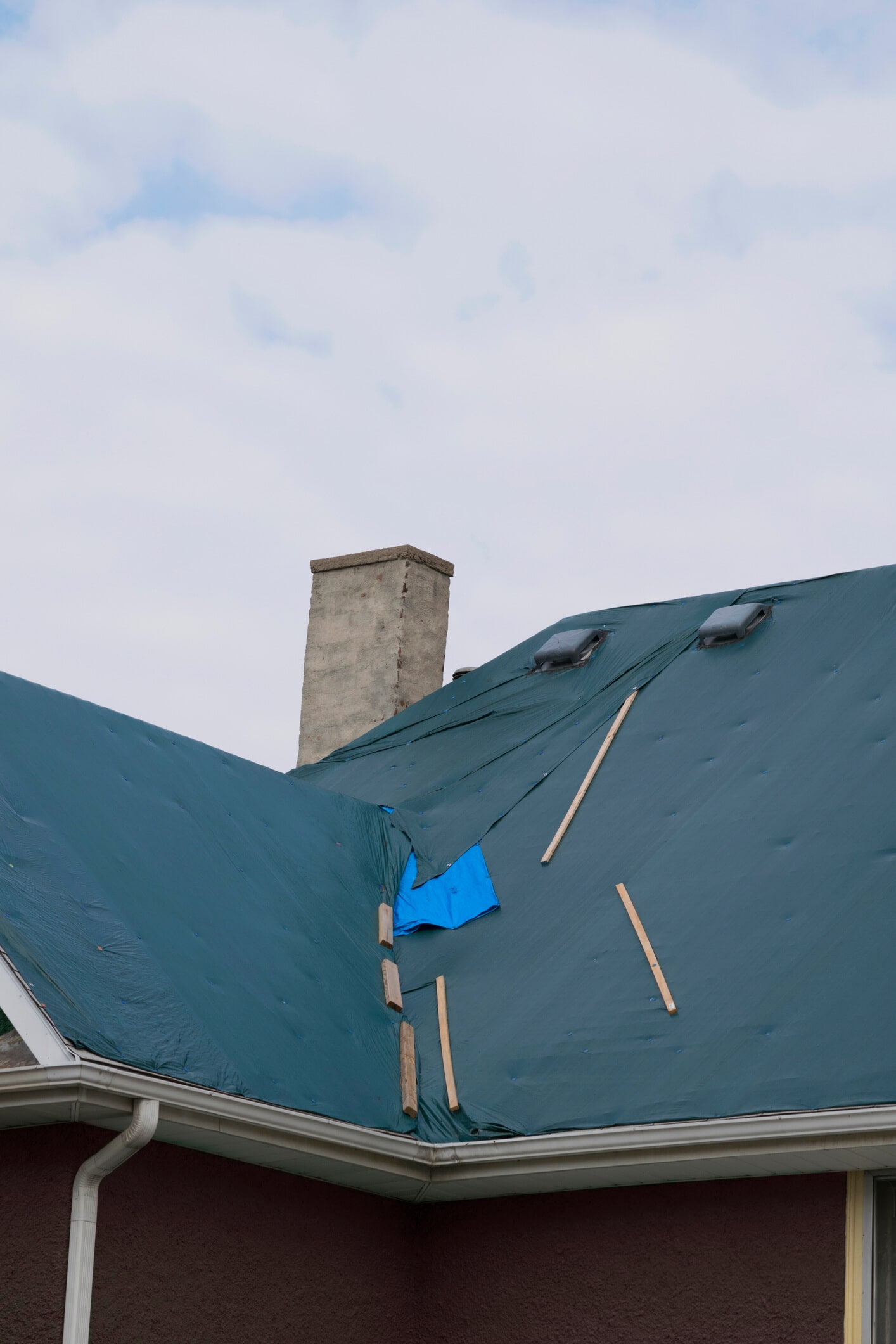 Blue tarp covering damaged roof with concrete chimney under cloudy sky